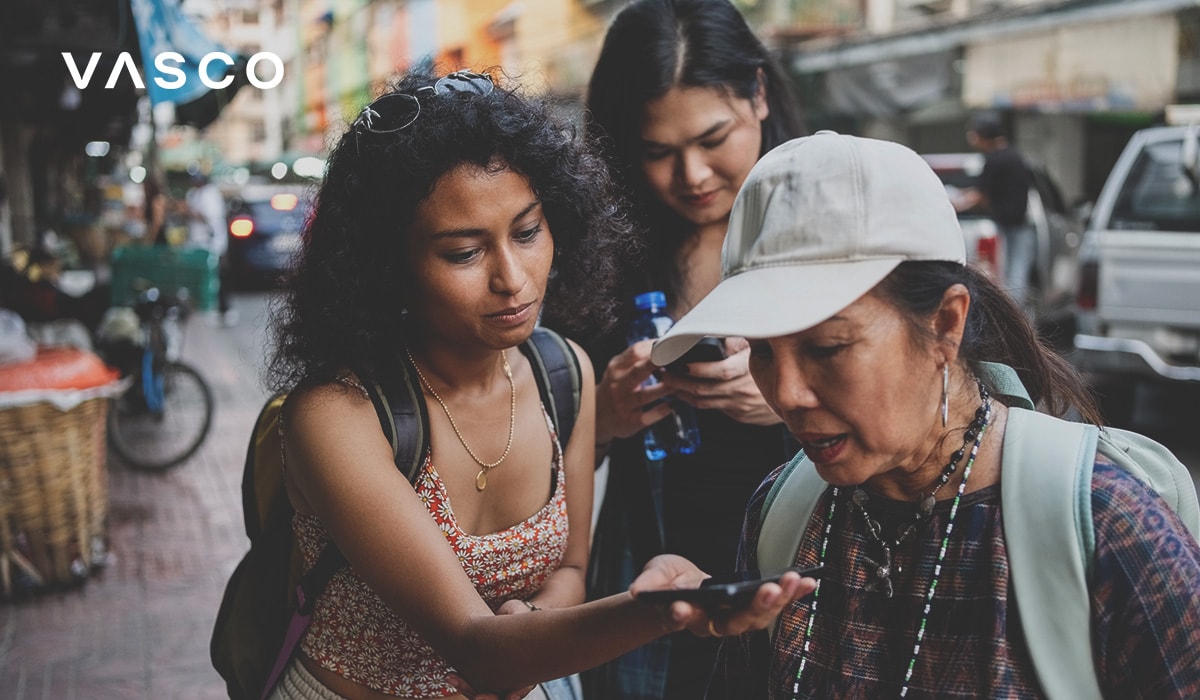 Un gruppo di tre persone in piedi su una strada affollata. Una persona tiene in mano uno smartphone, apparentemente dimostrando o utilizzando un'app di traduzione, mentre gli altri osservano con attenzione. L'ambientazione suggerisce un'interazione informale e autentica in un contesto multiculturale.