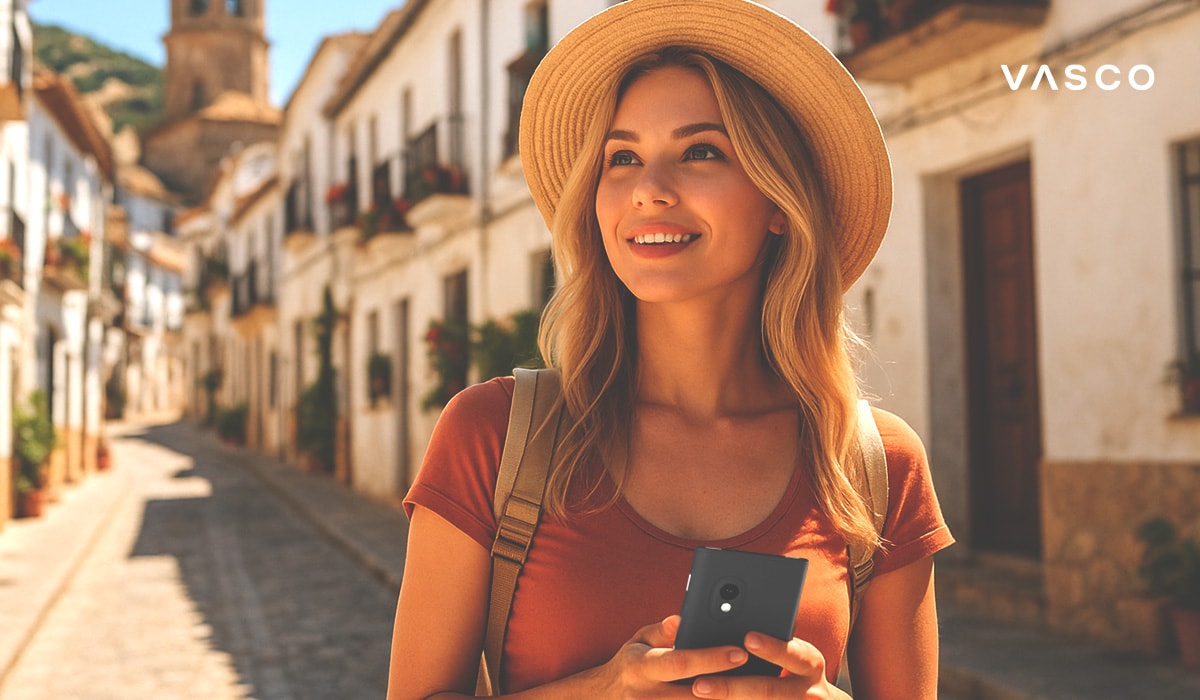 Turista sorridente che cammina per una stretta strada spagnola con edifici bianchi, tenendo un dispositivo di traduzione e indossando un cappello di paglia e uno zaino.
