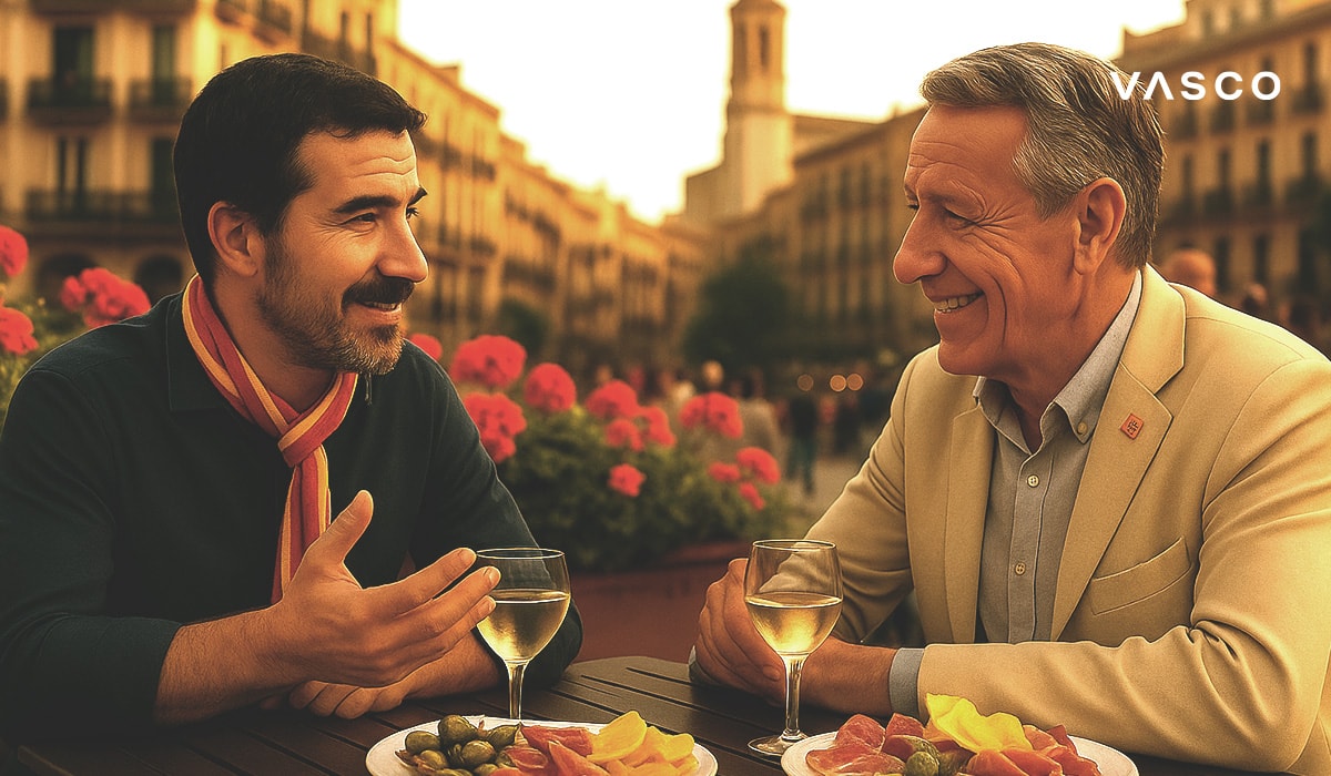 Due uomini che si godono drink e tapas in un caffè all’aperto in una piazza spagnola, immersi in conversazione, con fiori e architettura sullo sfondo.