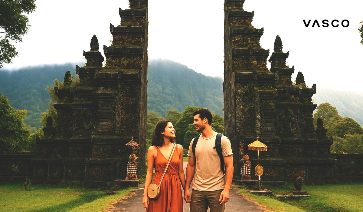 Una coppia si tiene per mano davanti a un tradizionale ingresso di un tempio balinese, con rigogliose montagne verdi sullo sfondo.