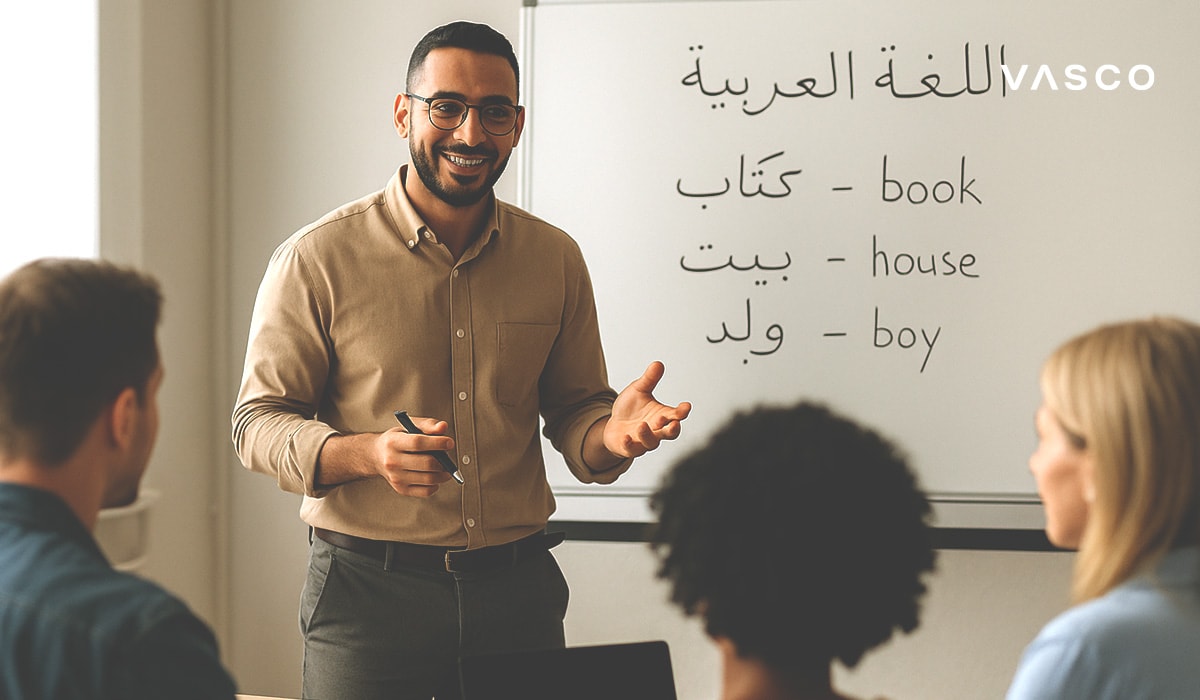 Insegnante che sorride durante una lezione di lingua araba, con parole arabe e relative traduzioni in inglese sulla lavagna.