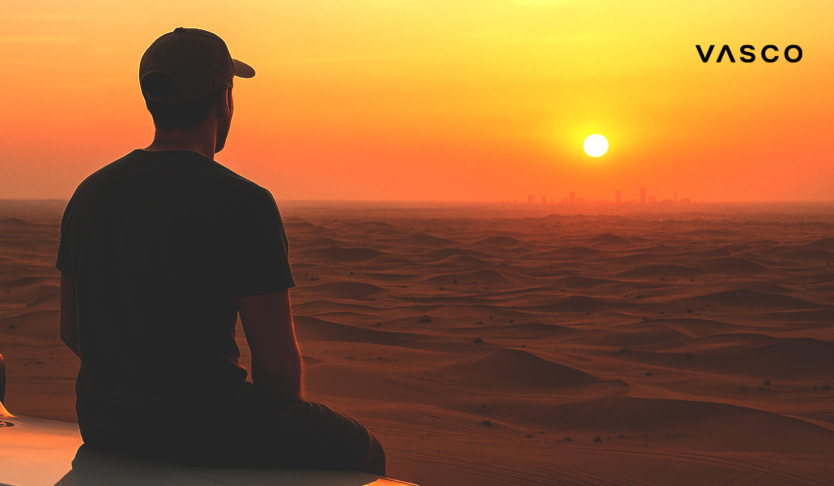 blog-01-uniwersal-arabic Uomo con un cappello seduto nel deserto che guarda il tramonto, con lo skyline di una città sullo sfondo.
