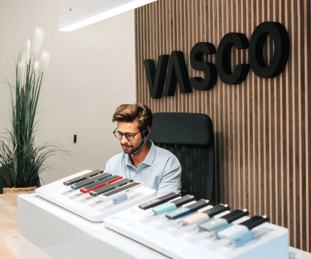 Un uomo del servizio clienti siede a una scrivania che espone diversi dispositivi Vasco Translator in vari colori, con un grande cartello 