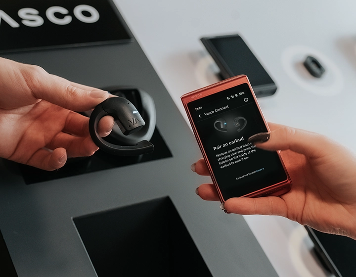 Vasco Translator E1 earbud and Vasco Translator Q1 shown together; the Q1 screen displays the earbud pairing interface, with the Vasco logo visible in the background.