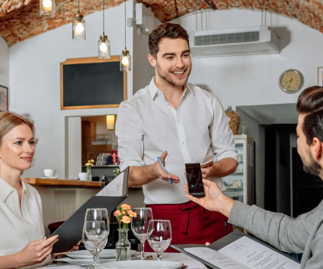 Un cameriere sorridente si trova al tavolo di un ristorante, mentre un cliente maschio seduto tiene in mano un traduttore. Una cliente donna siede vicino e tiene in mano un menu. Il tavolo è apparecchiato con bicchieri, piatti e un piccolo vaso di fiori.