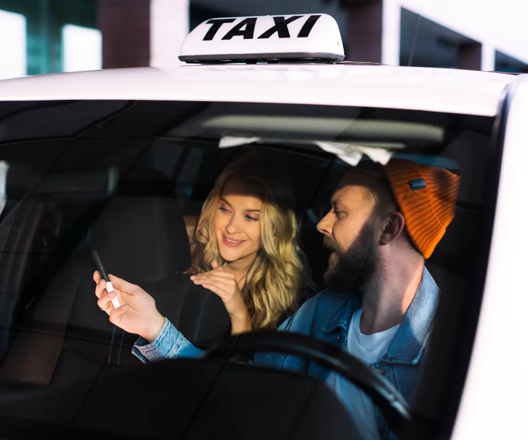 Un uomo siede al posto di guida di un taxi e mostra qualcosa a una donna sul sedile posteriore con un traduttore che tiene in mano. La donna sorride interessata. Sul tetto dell'auto è visibile l'insegna di un taxi.