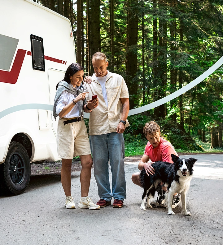 Due adulti in piedi accanto a un camper, concentrati su un Vasco Translator V4. Accanto a loro, il figlio accarezza dolcemente un cane, in una tranquilla scena di viaggio in famiglia.
