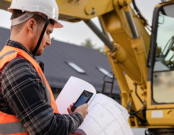 Un uomo con il casco e indosso abiti da lavoro mentre scatta una foto dei progetti di costruzione utilizzando Vasco Translator V4, con un escavatore visibile sullo sfondo.