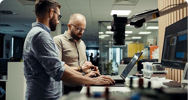 Due uomini con la barba e gli occhiali stanno lavorando insieme a una scrivania in piedi in un moderno ufficio o laboratorio. Uno dei due, con una camicia azzurra, sta usando un computer portatile, mentre l’altro, con una camicia color cuoio, gli sta accanto indicando con un gesto un piccolo componente elettronico che tiene in mano. La scrivania è dotata di un grande monitor, vari strumenti elettronici e una telecamera montata su un braccio regolabile. Sullo sfondo si vede uno spazio di lavoro luminoso e open space con pareti divisorie in vetro. 