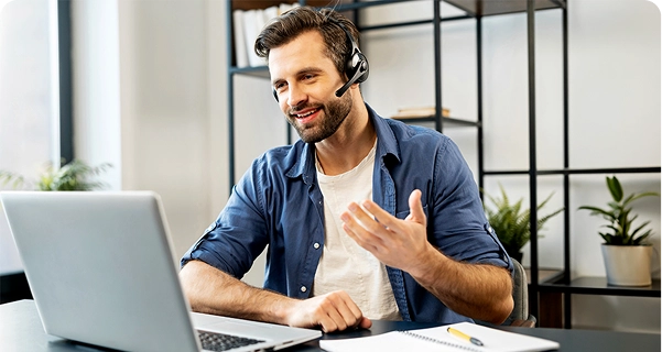 Un uomo dall'aspetto cordiale, con la barba e i capelli scuri, è seduto a una scrivania e indossa delle cuffie professionali dotate di microfono. Sta sorridendo e gesticolando con una mano mentre guarda lo schermo di un portatile; sembra che sia nel bel mezzo di una videochiamata o che stia fornendo assistenza tecnica. Indossa una camicia blu abbottonata sopra una maglietta bianca. Sulla scrivania davanti a lui ci sono un laptop, un taccuino e una penna gialla. Lo sfondo è un ufficio luminoso e moderno con una scaffalatura minimalista in metallo nero e piante in vaso verdi. 