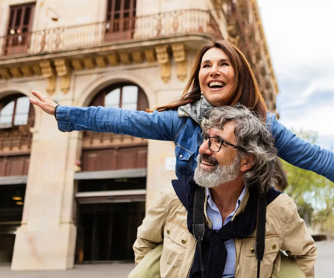 Un anziano allegro porta una donna sulle spalle per le strade di una città europea; entrambi sorridono a trentadue denti mentre lei indica con il braccio alzato verso l’orizzonte. Lui indossa occhiali, una giacca color cuoio e ha con sé una macchina fotografica, mentre lei indossa una giacca di jeans e una sciarpa. 