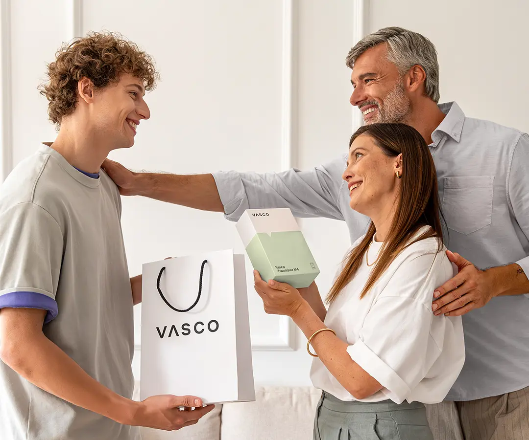 Una scena commovente in cui tre persone sorridono in piedi una accanto all’altra. A sinistra, un giovane dai capelli ricci tiene in mano una borsa della spesa bianca con la scritta «VASCO». Al centro, una donna tiene in mano una scatola bianca e verde chiaro con l’etichetta «Vasco Translator M4», guardando felice il giovane. Dietro di lei, un uomo dai capelli brizzolati sorride calorosamente, con una mano sulla spalla della donna e l'altra su quella del giovane. Si trovano in una stanza luminosa con pareti dai colori neutri e un divano sullo sfondo. 