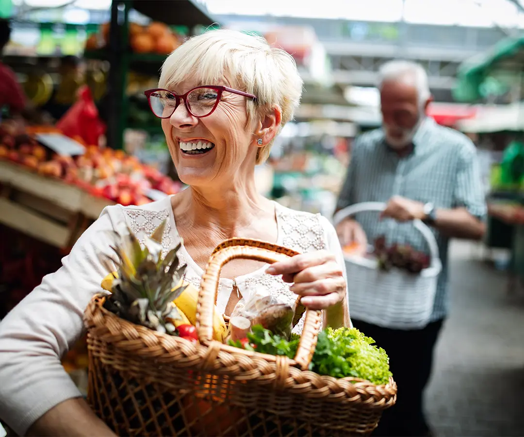 Una signora anziana sorridente, con i capelli biondi corti e occhiali rossi, che porta un cesto di vimini pieno di frutta e verdura fresca in un mercato all'aperto. Sullo sfondo sfocato, un'altra persona sta facendo la spesa tra le bancarelle di prodotti ortofrutticoli.