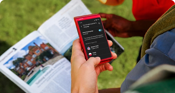 Una persona utilizza un Vasco Translator Q1 rosso per scattare una foto di un giornale. Lo schermo mostra il menù di selezione della lingua per la traduzione del testo rilevato automaticamente dal giornale.